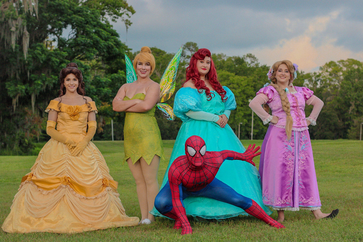 Five people dressed as Disney princesses and Spider-Man pose together on a grassy lawn with trees in the background. From left to right: Belle in a yellow gown, a fairy character in a green dress, Ariel in a turquoise gown, Spider-Man in his red and blue suit, and Princess Peach in a pink gown.