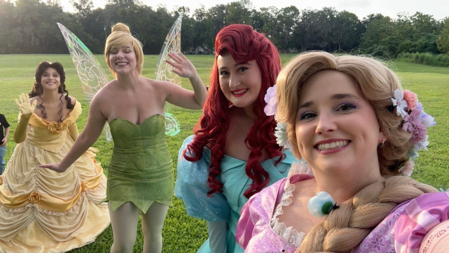 Four women posing outdoors in Disney princess costumes on a grassy field, including Belle in yellow, Tinker Bell in green, Ariel in light blue, and Rapunzel in pink with a braid and flower hair accessory.