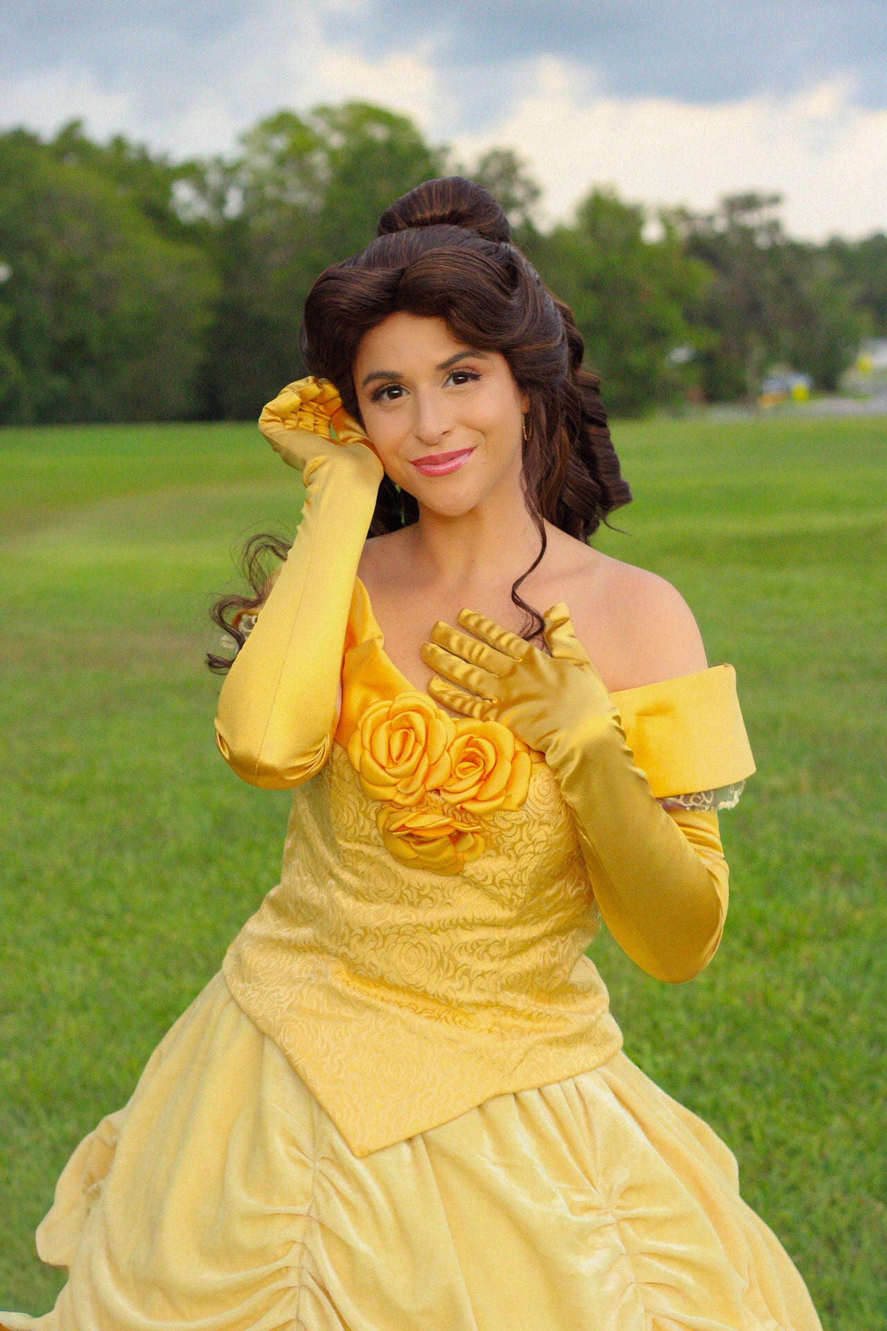 A woman posing outdoors in a yellow Belle de Ville cosplay costume, featuring a yellow off-the-shoulder dress with rose details on the bodice, matching yellow gloves, and a styled updo.