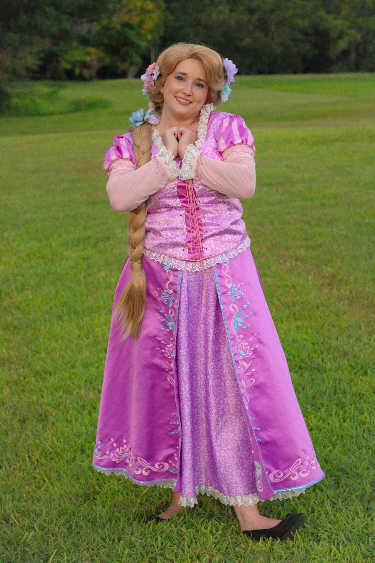 A woman wearing a pink and purple Rapunzel-style costume stands barefoot on a green grassy lawn, with her hands together in a prayer pose. The costume features a shiny pink bodice, a long purple skirt with embroidered details, and a long blonde wig with blue flowers.