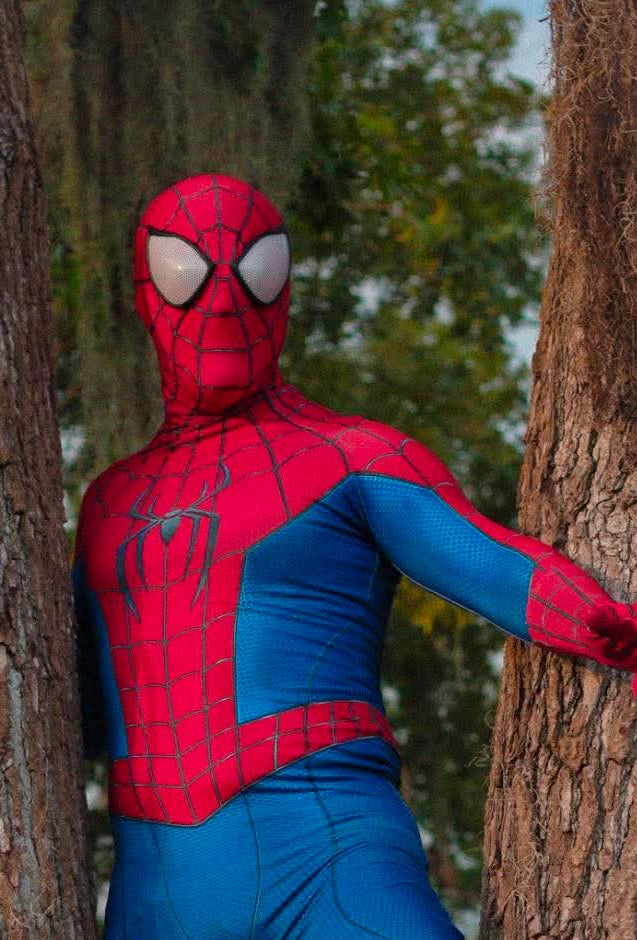 A person wearing a full Spider-Man costume posed between two large tree trunks outdoors. The costume is red and blue with black web details and a spider emblem on the chest, and the person's face is covered by a red mask with white eyes.