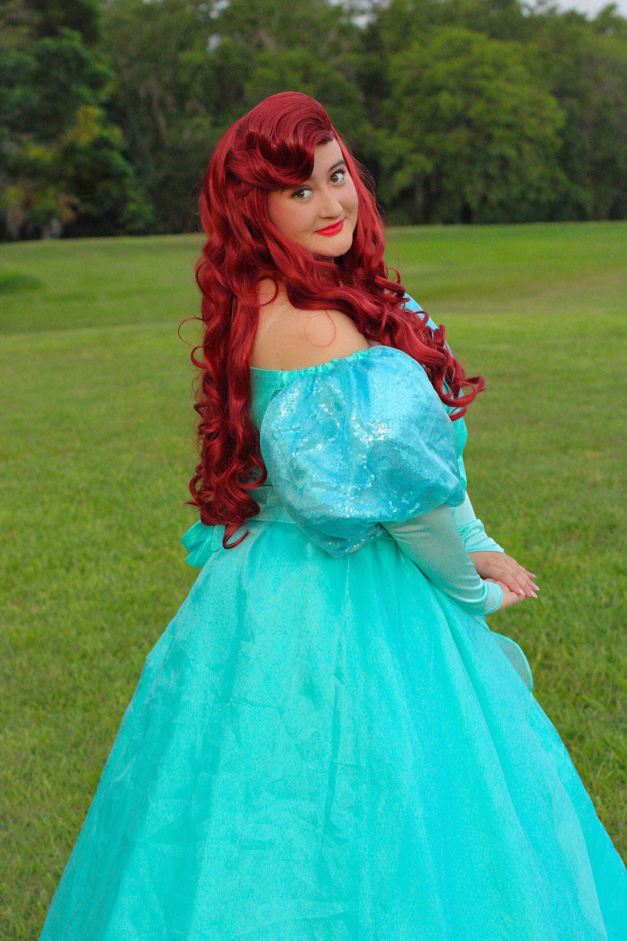 A woman outdoors in a grassy park wearing a turquoise off-the-shoulder ballgown costume with long red hair and red lipstick.